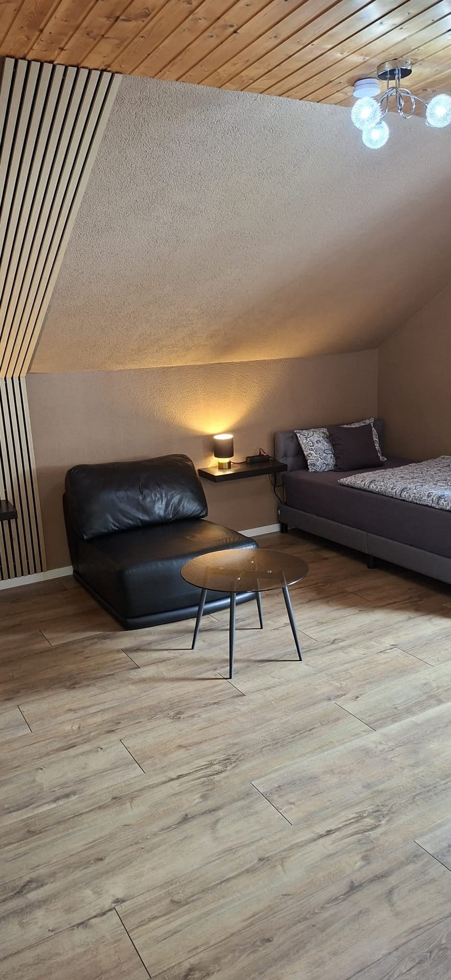Wohn- und Schlafbereich der Wohnung B mit Holzdecke, Lamellenwand, schwarzem Ledersessel und Bett