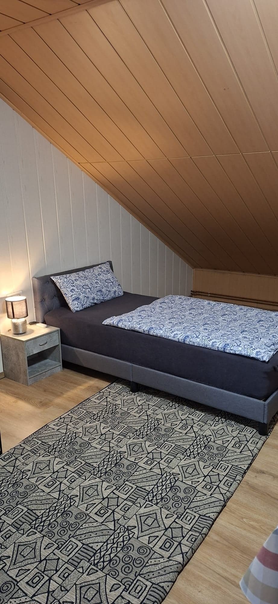 Schlafzimmer der Wohnung B mit Einzelbett unter der hellen Holzdecke und gemustertem Teppich